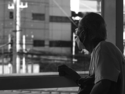 A senior man wearing a face mask looks thoughtfully out of a window in black and white.