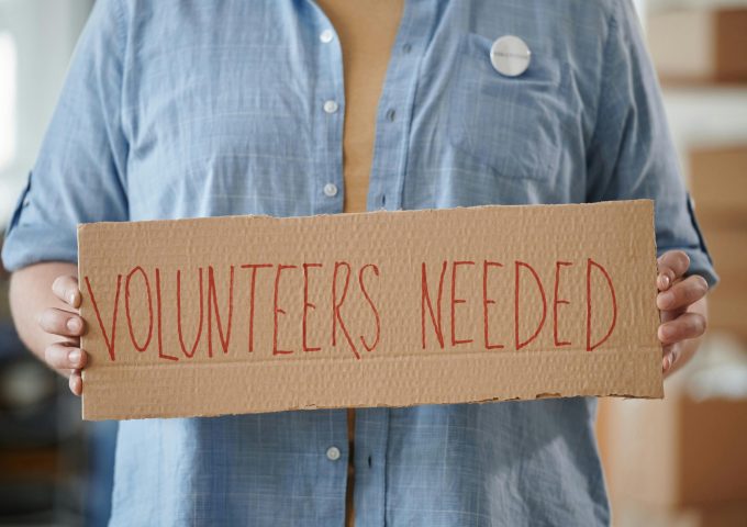 Person holding a sign reading 'Volunteers Needed' to encourage community support.